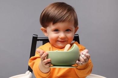 Cute little kid eating healthy baby food from bowl in high chair on light grey background