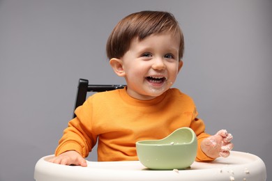 Cute little kid eating healthy baby food from bowl in high chair on light grey background