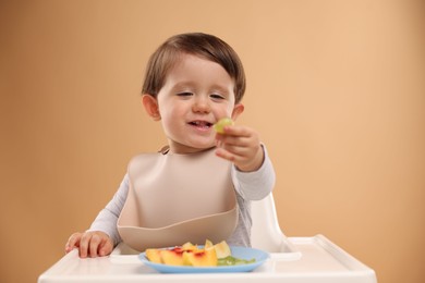 Photo of Healthy baby food. Cute little kid eating fruits in high chair on beige background