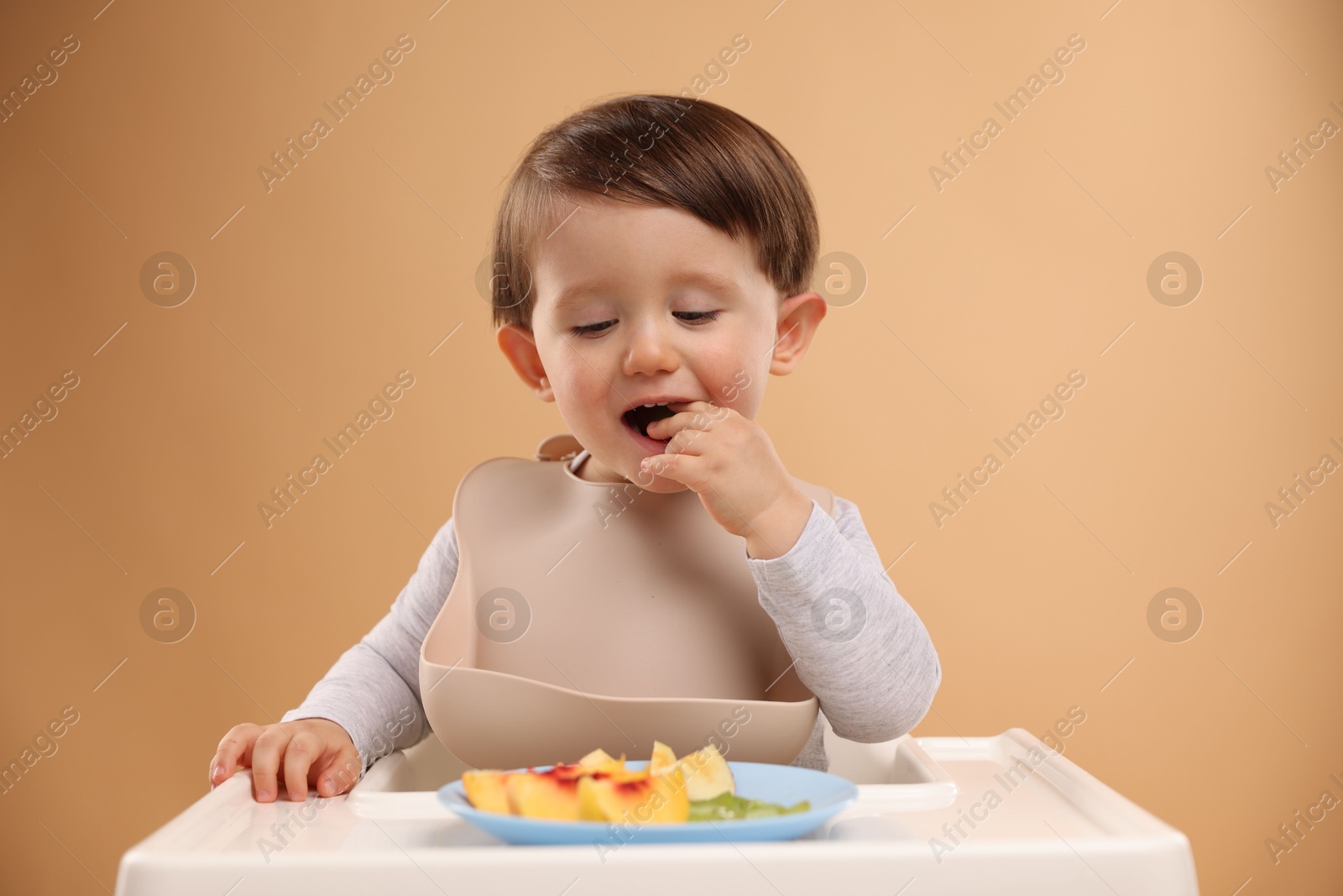 Photo of Healthy baby food. Cute little kid eating fruits in high chair on beige background