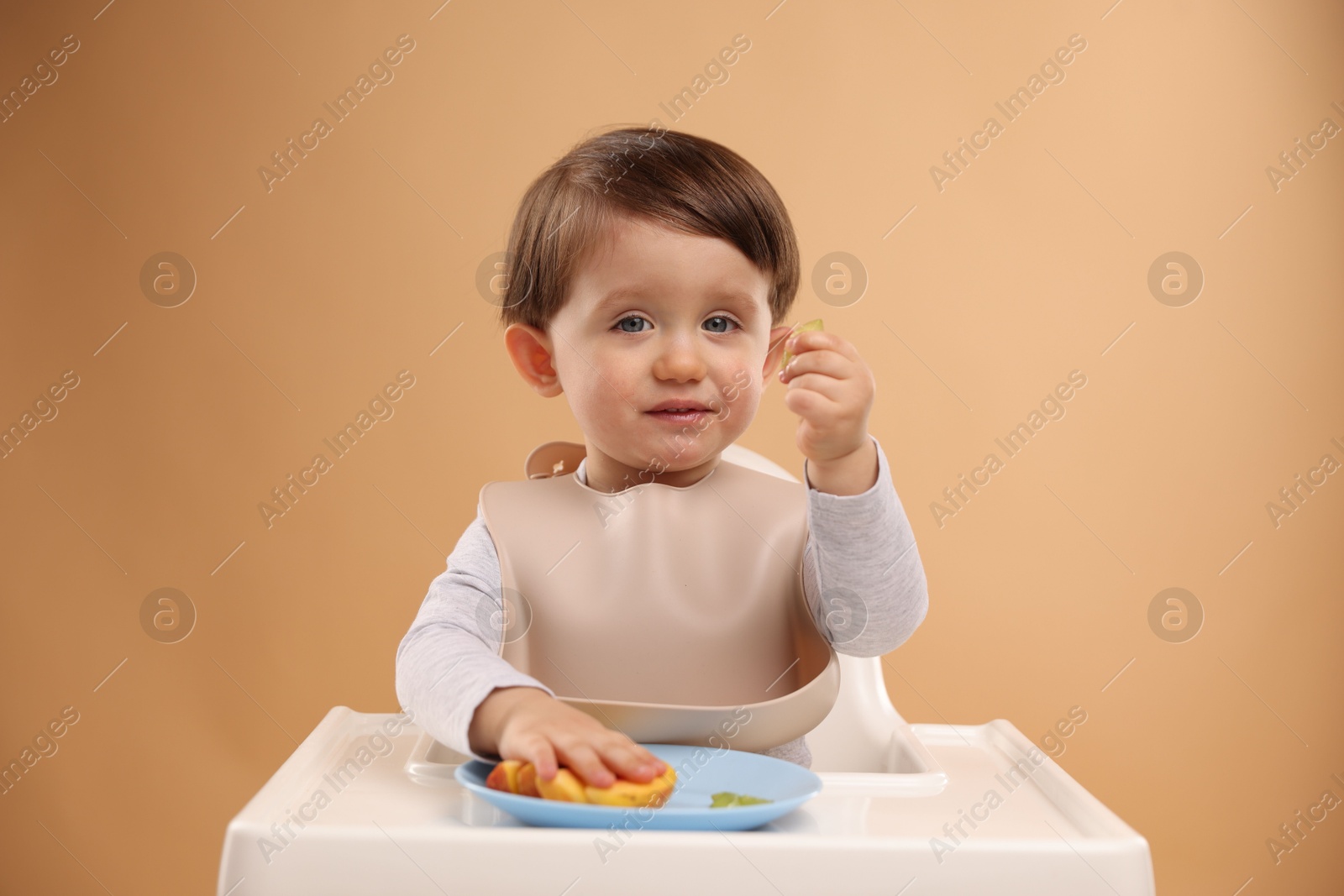 Photo of Healthy baby food. Cute little kid eating fruits in high chair on beige background