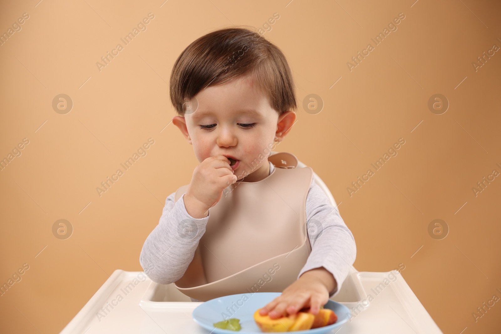 Photo of Healthy baby food. Cute little kid eating fruits in high chair on beige background