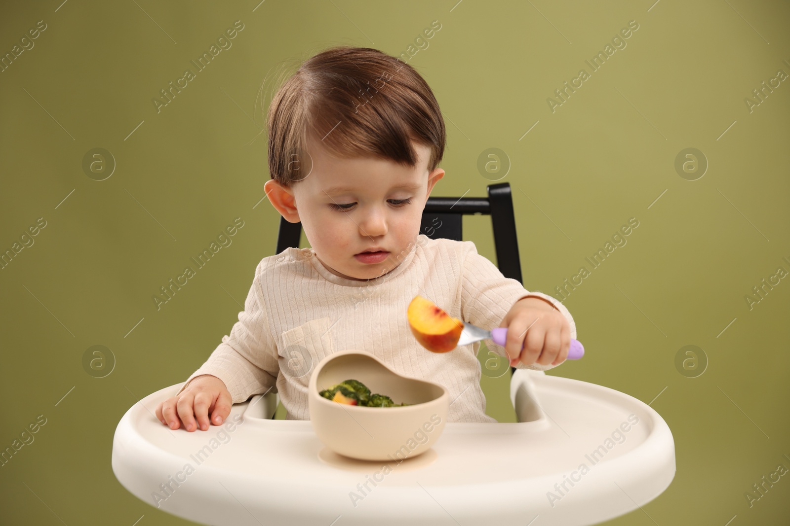 Photo of Cute little baby eating healthy food from bowl in high chair on olive background