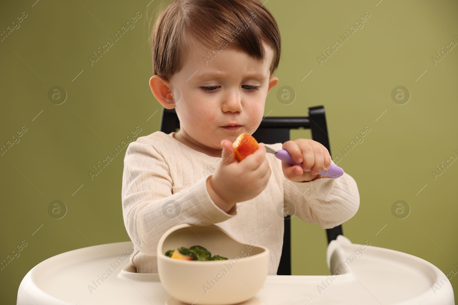 Photo of Cute little baby eating healthy food from bowl in high chair on olive background