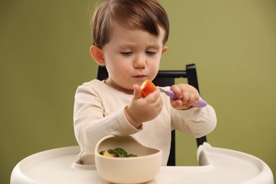 Cute little baby eating healthy food from bowl in high chair on olive background