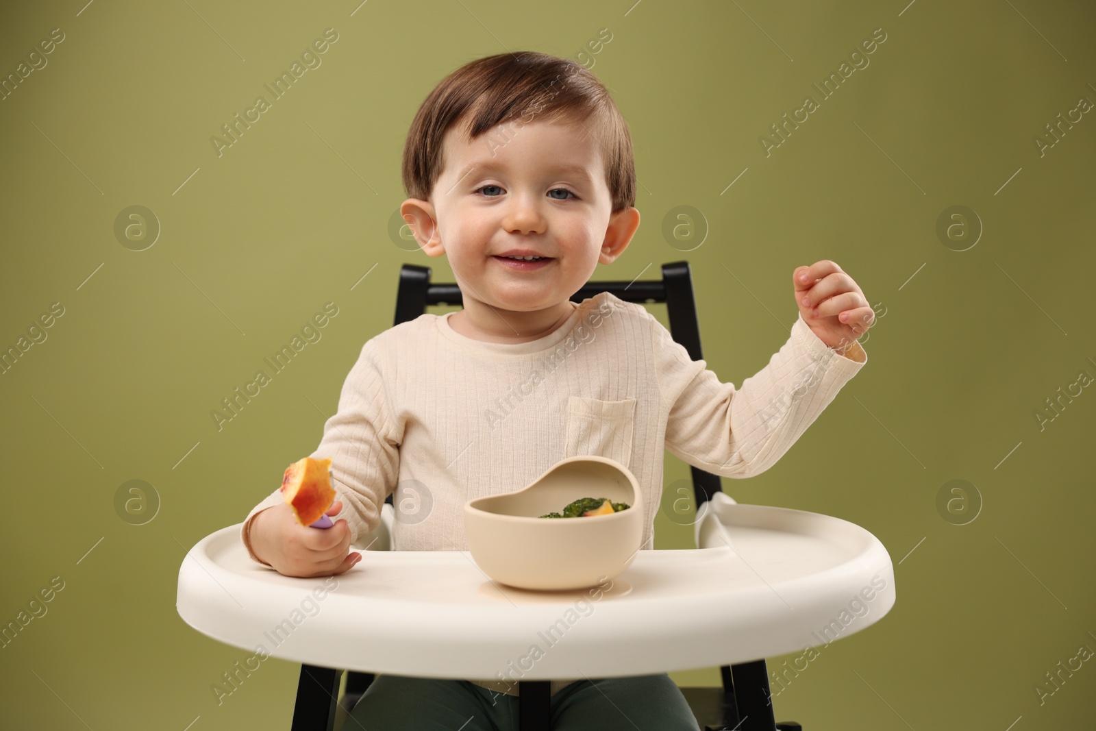 Photo of Cute little baby eating healthy food from bowl in high chair on olive background