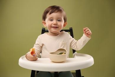 Photo of Cute little baby eating healthy food from bowl in high chair on olive background