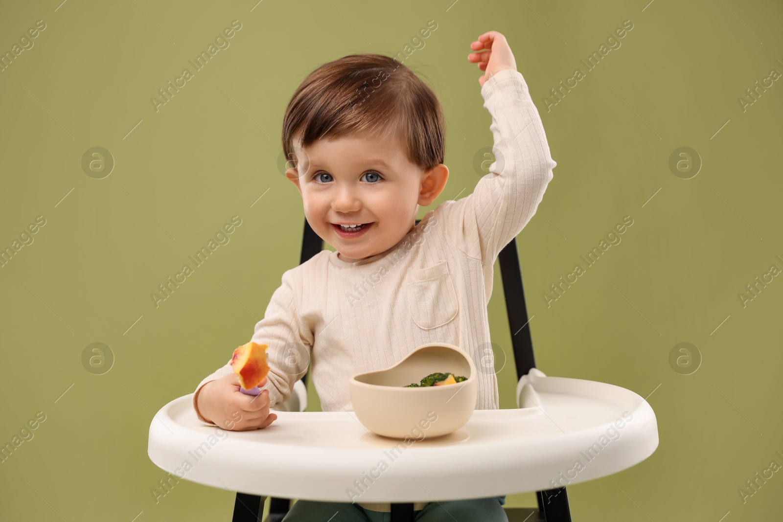 Photo of Cute little baby eating healthy food from bowl in high chair on olive background