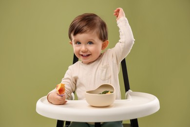 Cute little baby eating healthy food from bowl in high chair on olive background