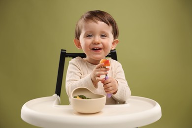 Photo of Cute little baby eating healthy food from bowl in high chair on olive background