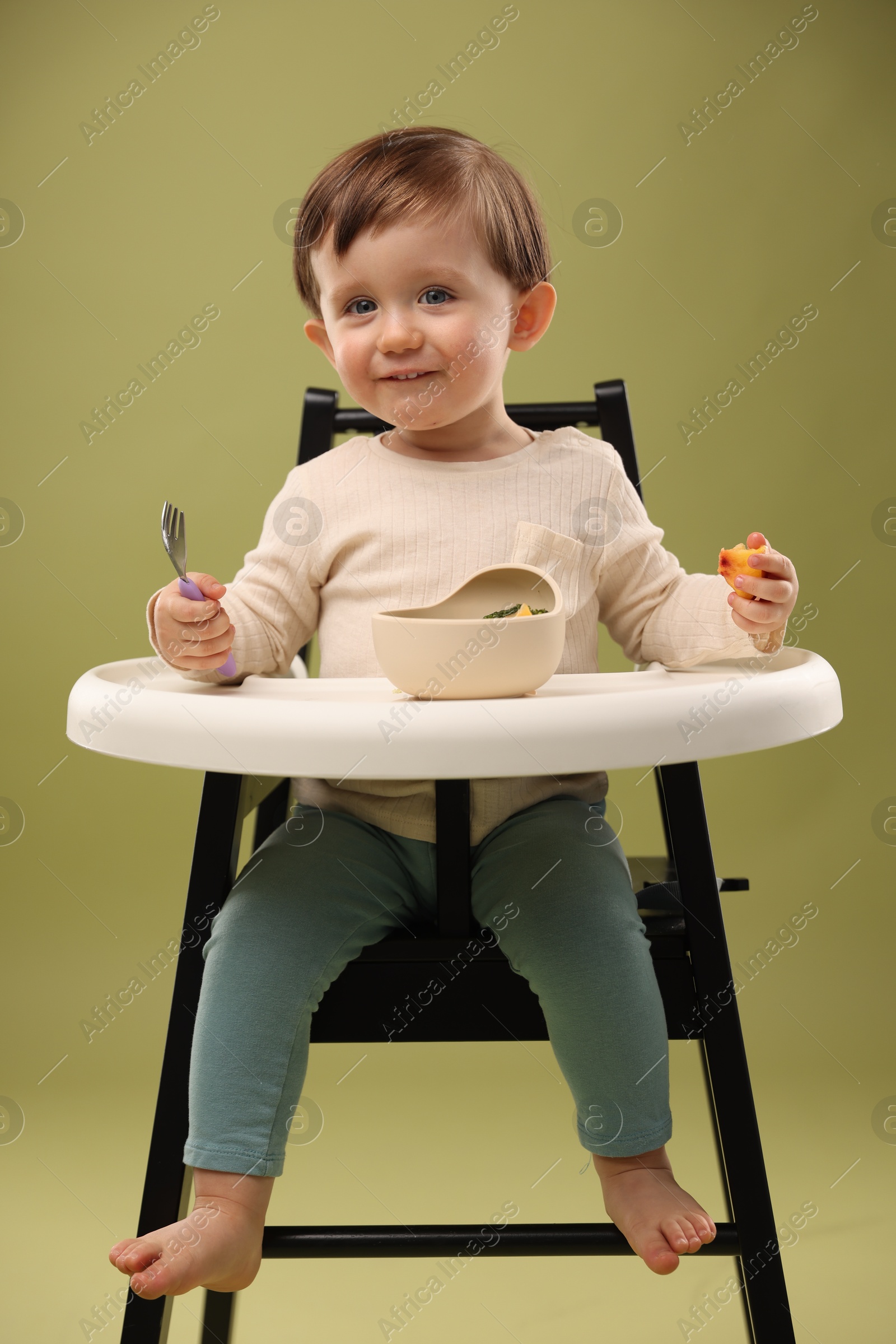 Photo of Cute little baby eating healthy food from bowl in high chair on olive background