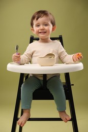 Photo of Cute little baby eating healthy food from bowl in high chair on olive background