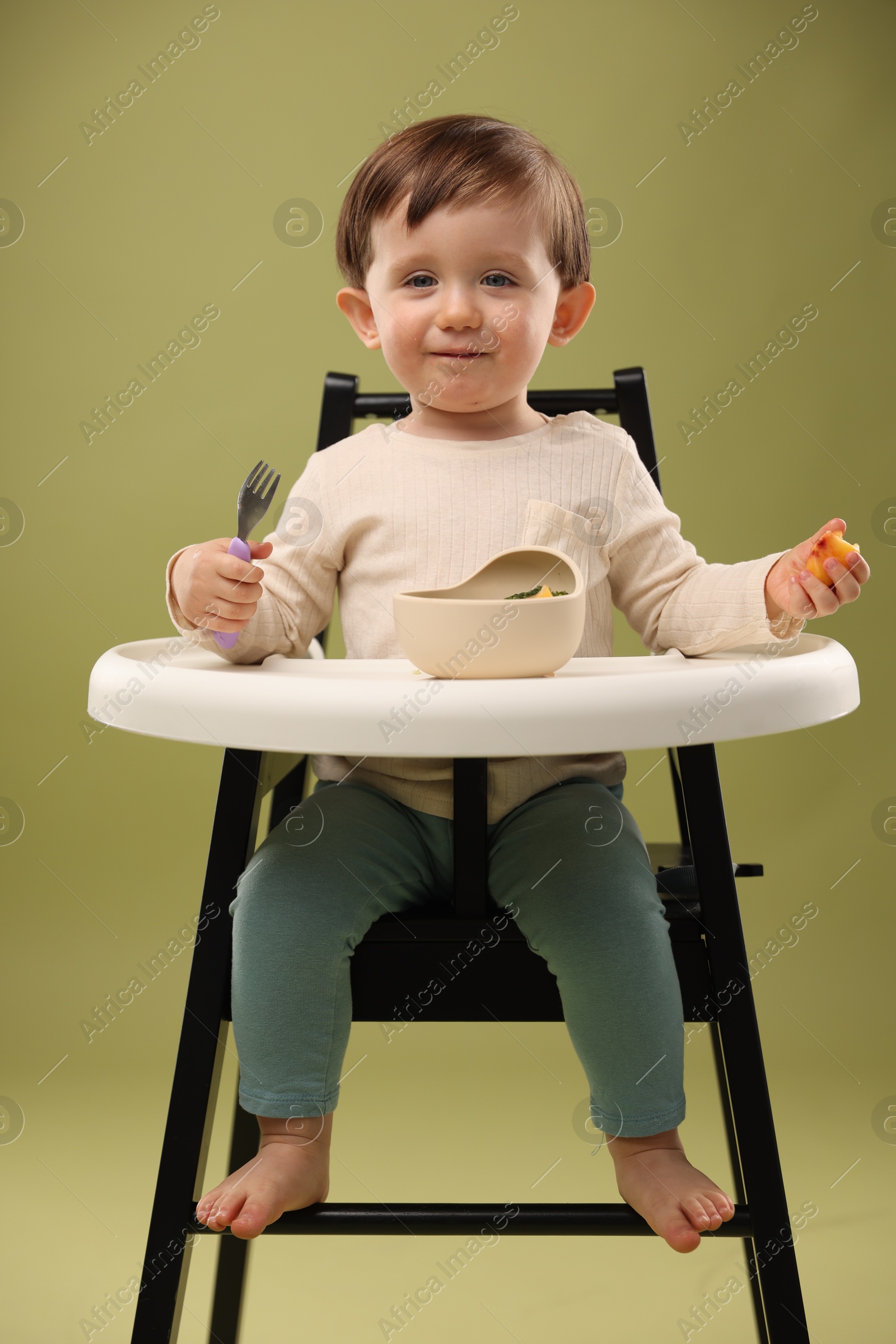 Photo of Cute little baby eating healthy food from bowl in high chair on olive background