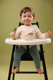 Cute little baby eating healthy food from bowl in high chair on olive background