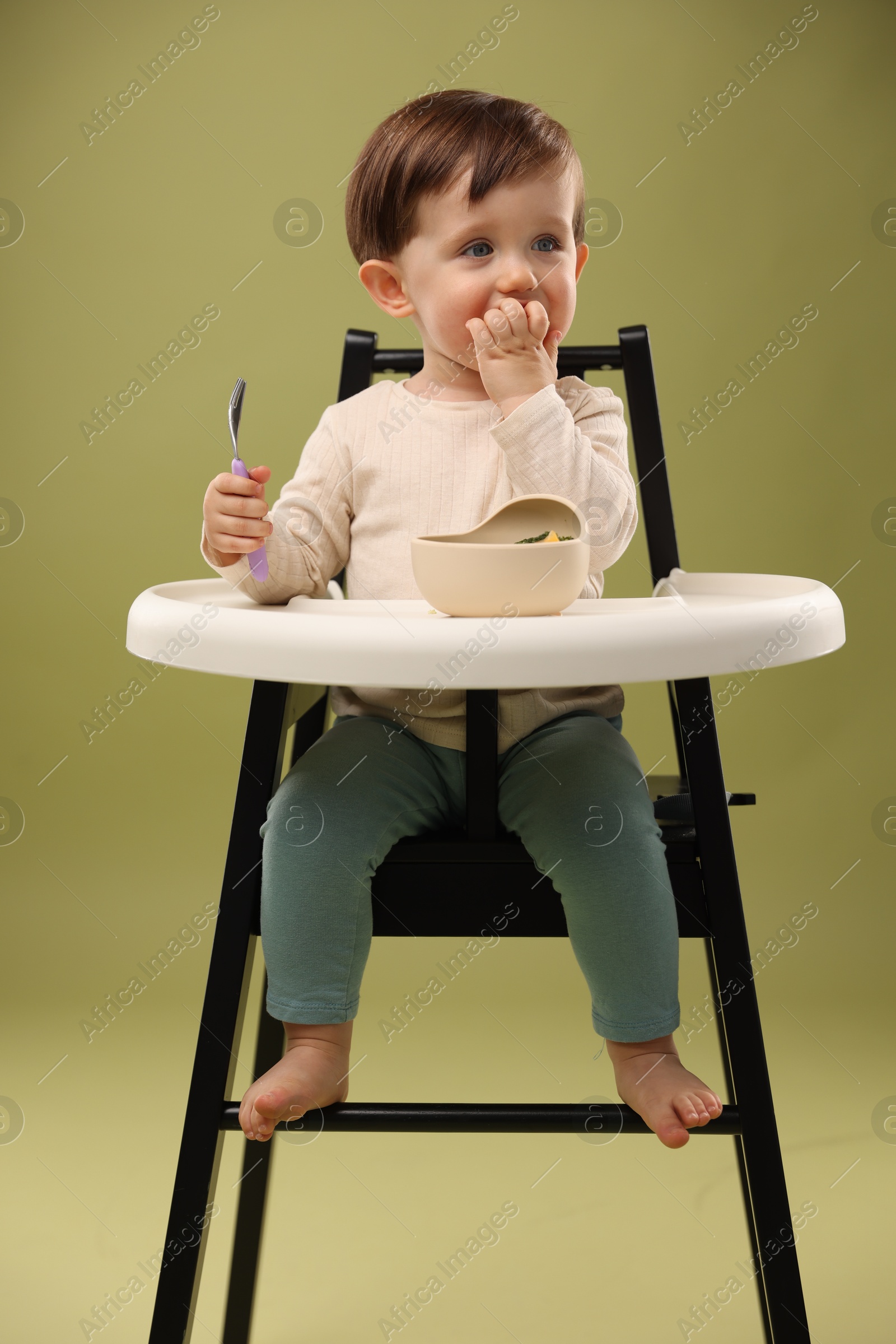 Photo of Cute little baby eating healthy food from bowl in high chair on olive background