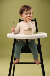 Cute little baby eating healthy food from bowl in high chair on olive background