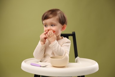 Photo of Cute little baby eating healthy food from bowl in high chair on olive background