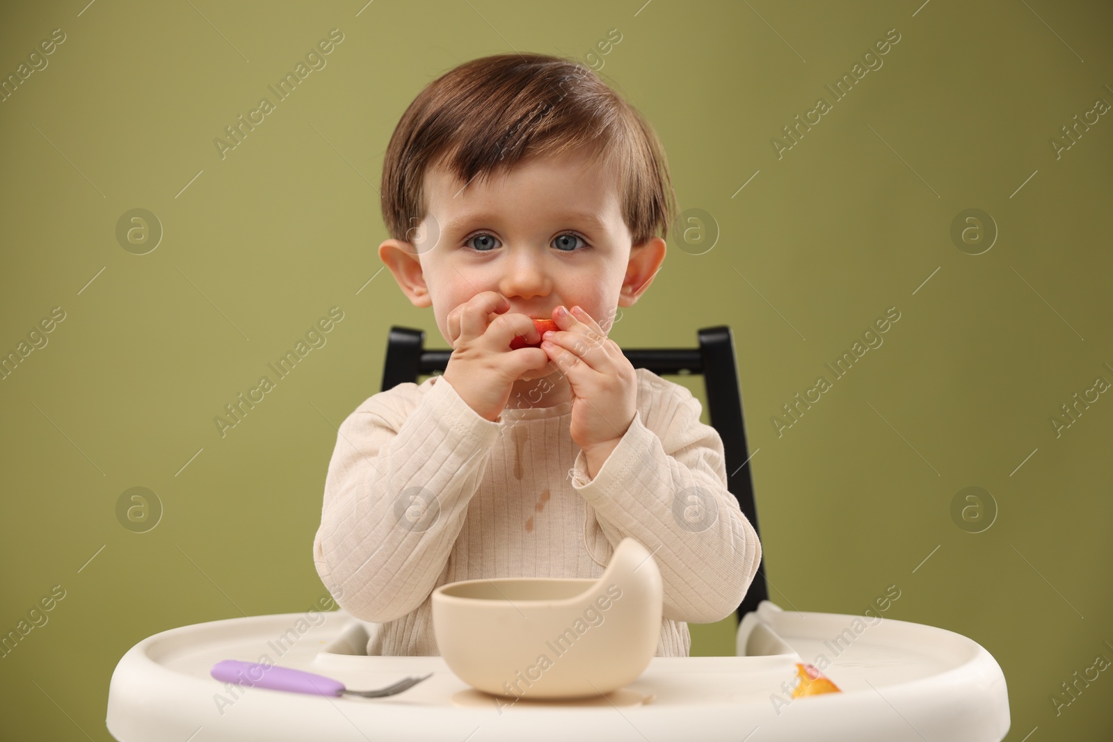 Photo of Cute little baby eating healthy food from bowl in high chair on olive background