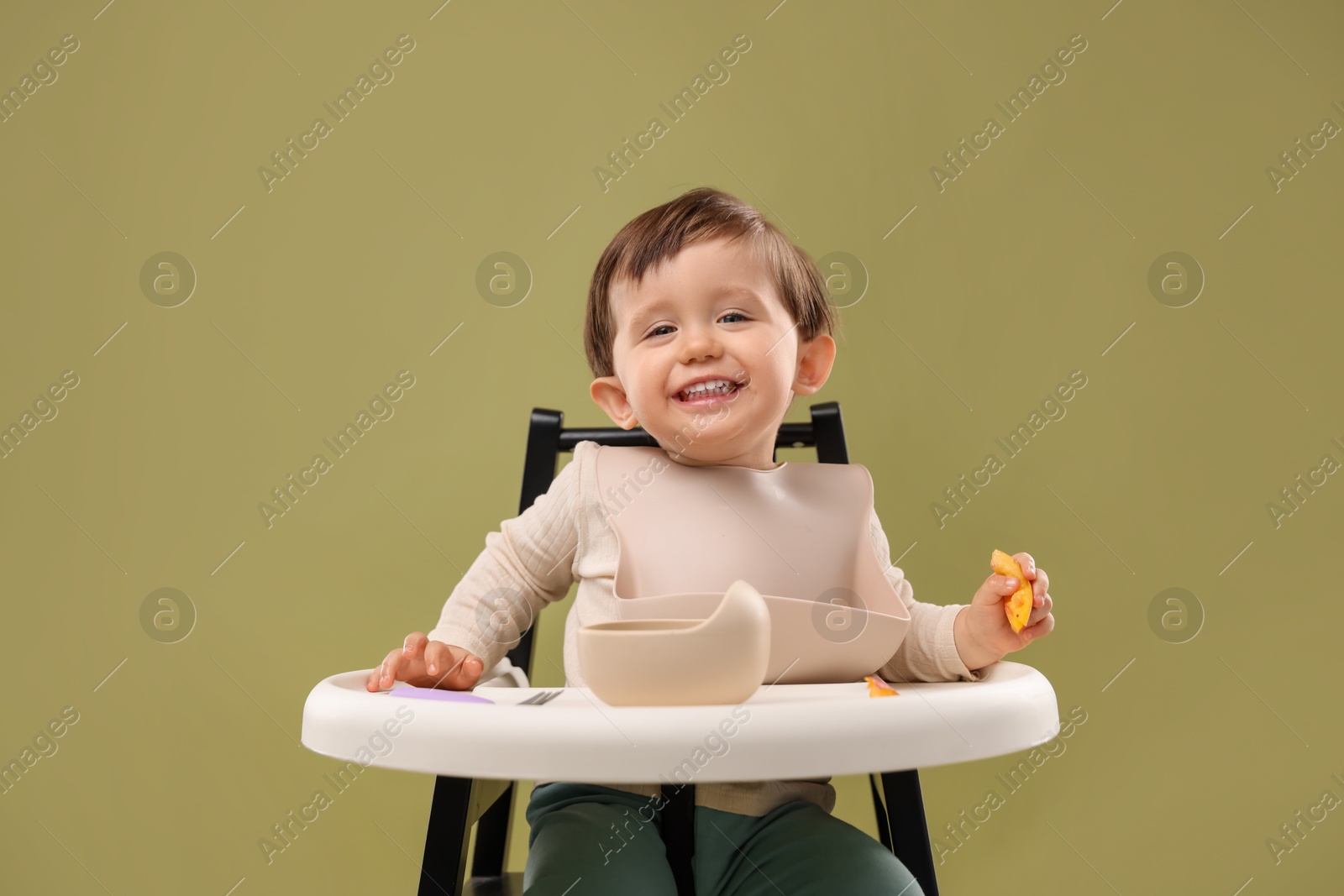 Photo of Cute little baby eating healthy food from bowl in high chair on olive background