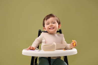 Cute little baby eating healthy food from bowl in high chair on olive background