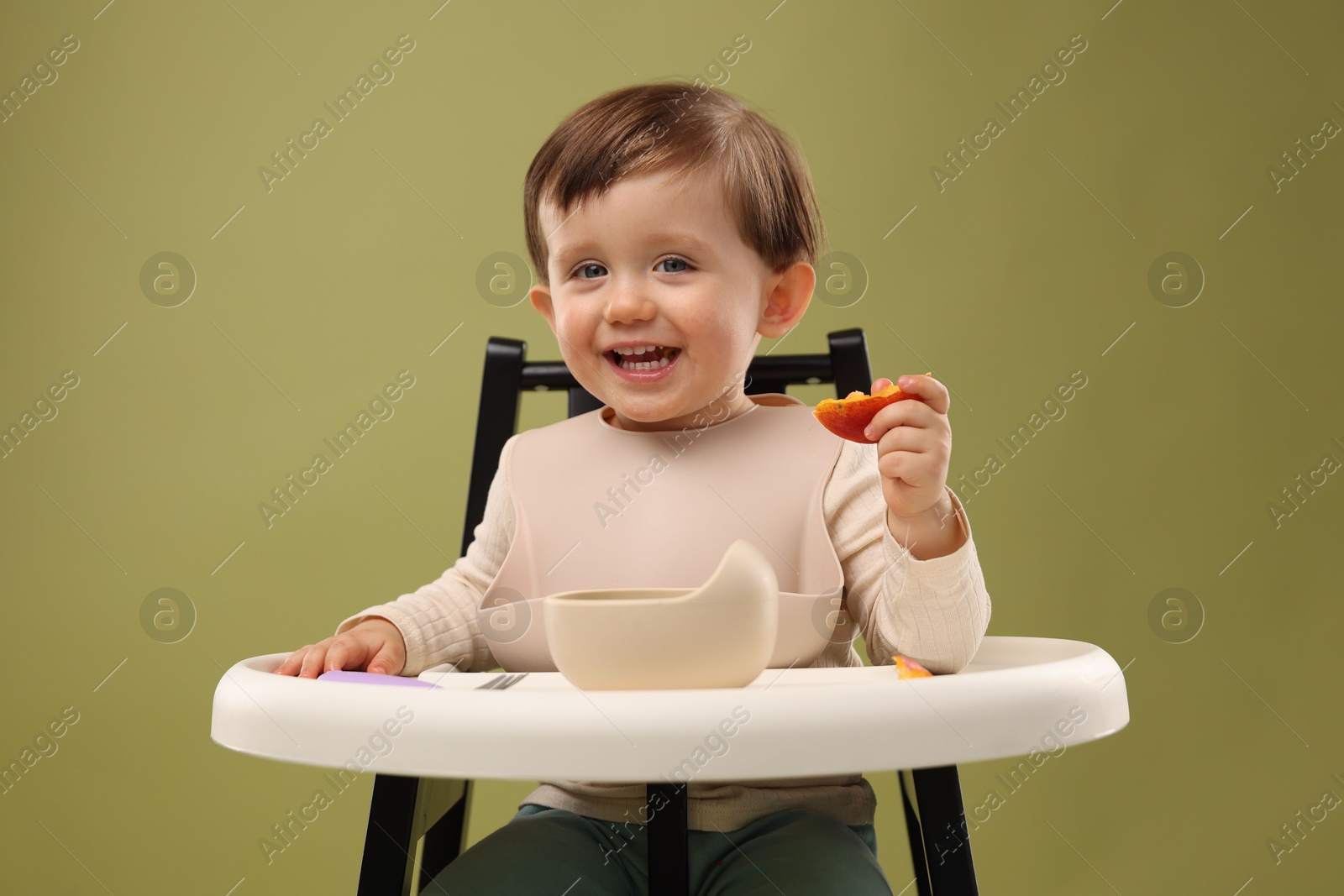 Photo of Cute little baby eating healthy food from bowl in high chair on olive background