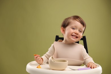 Cute little baby eating healthy food from bowl in high chair on olive background, space for text