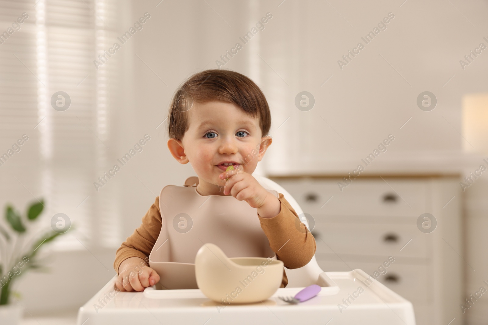 Photo of Cute little baby eating healthy food from bowl in high chair at home