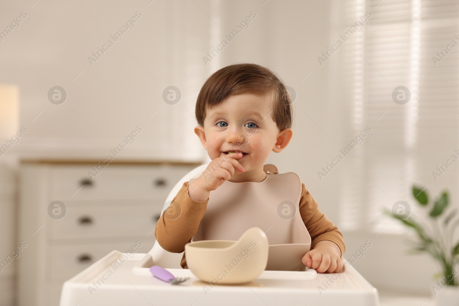 Photo of Cute little baby eating healthy food from bowl in high chair at home