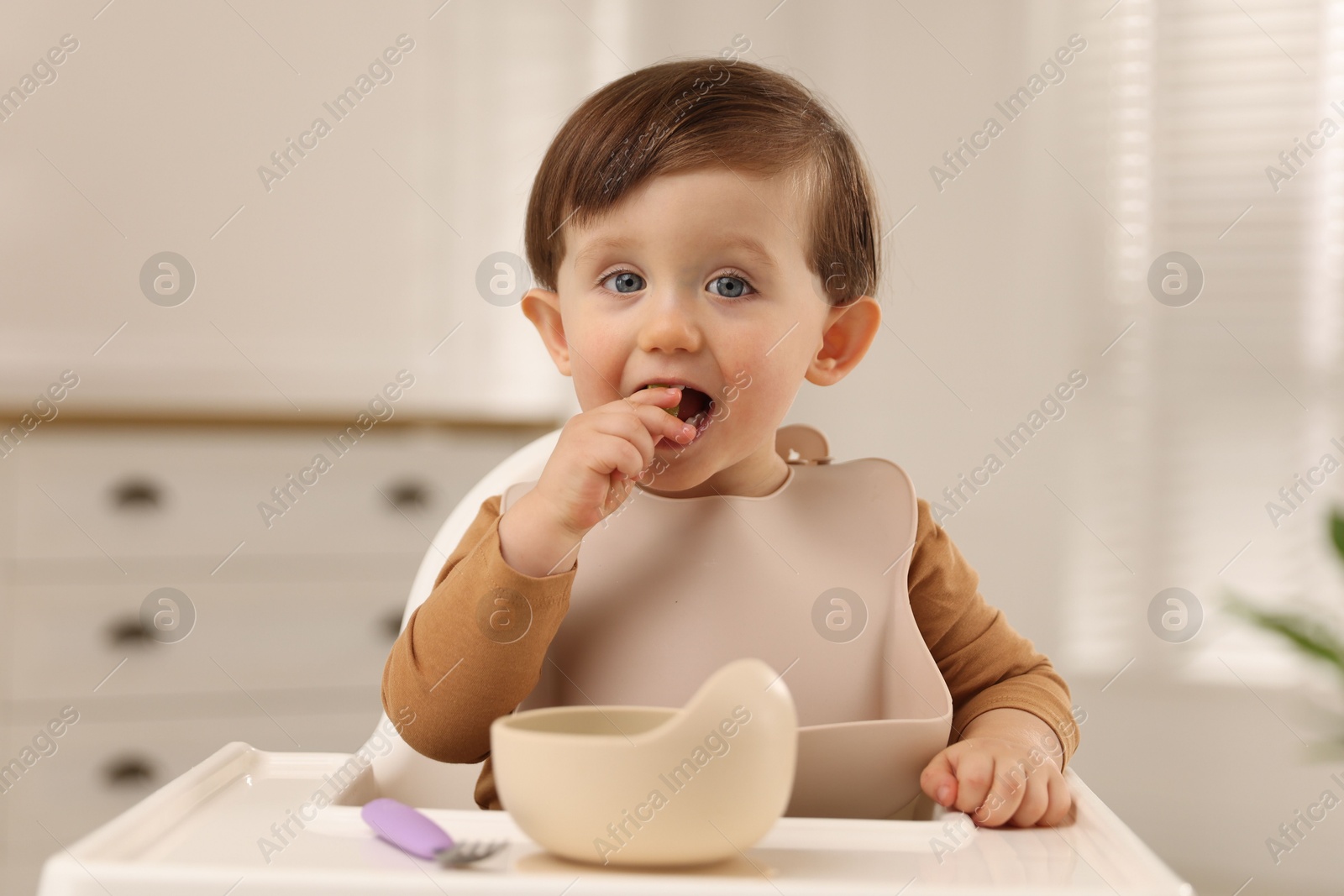 Photo of Cute little baby eating healthy food from bowl in high chair at home