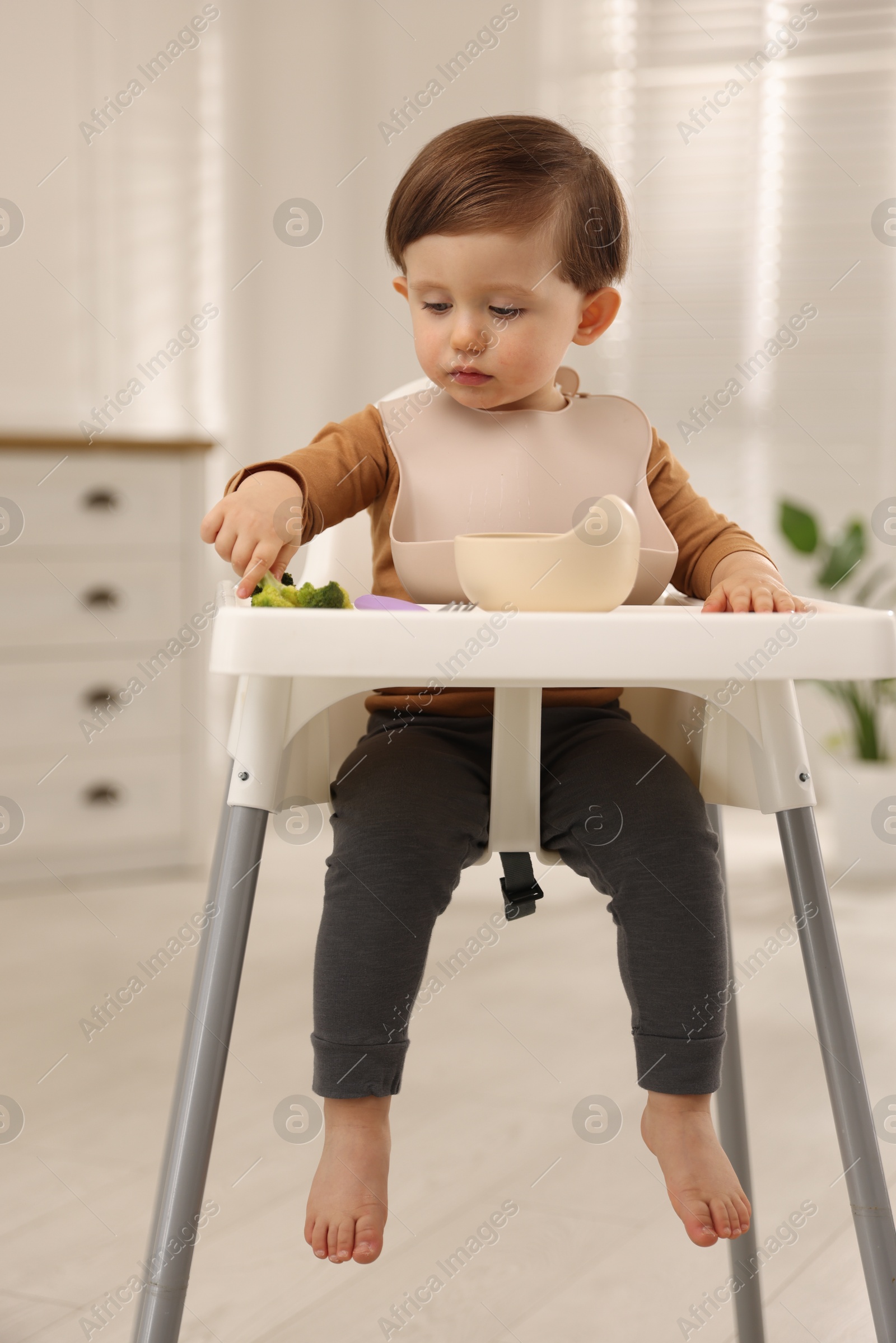 Photo of Cute little baby eating healthy food from bowl in high chair at home