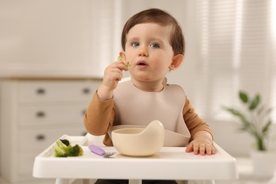 Cute little baby eating healthy food from bowl in high chair at home