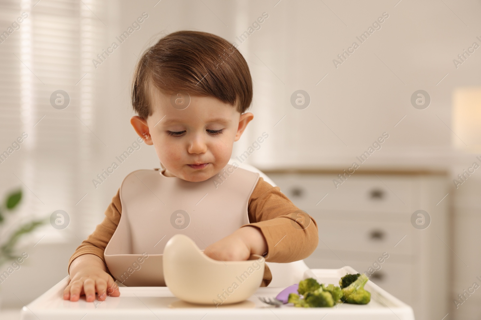 Photo of Cute little baby eating healthy food from bowl in high chair at home