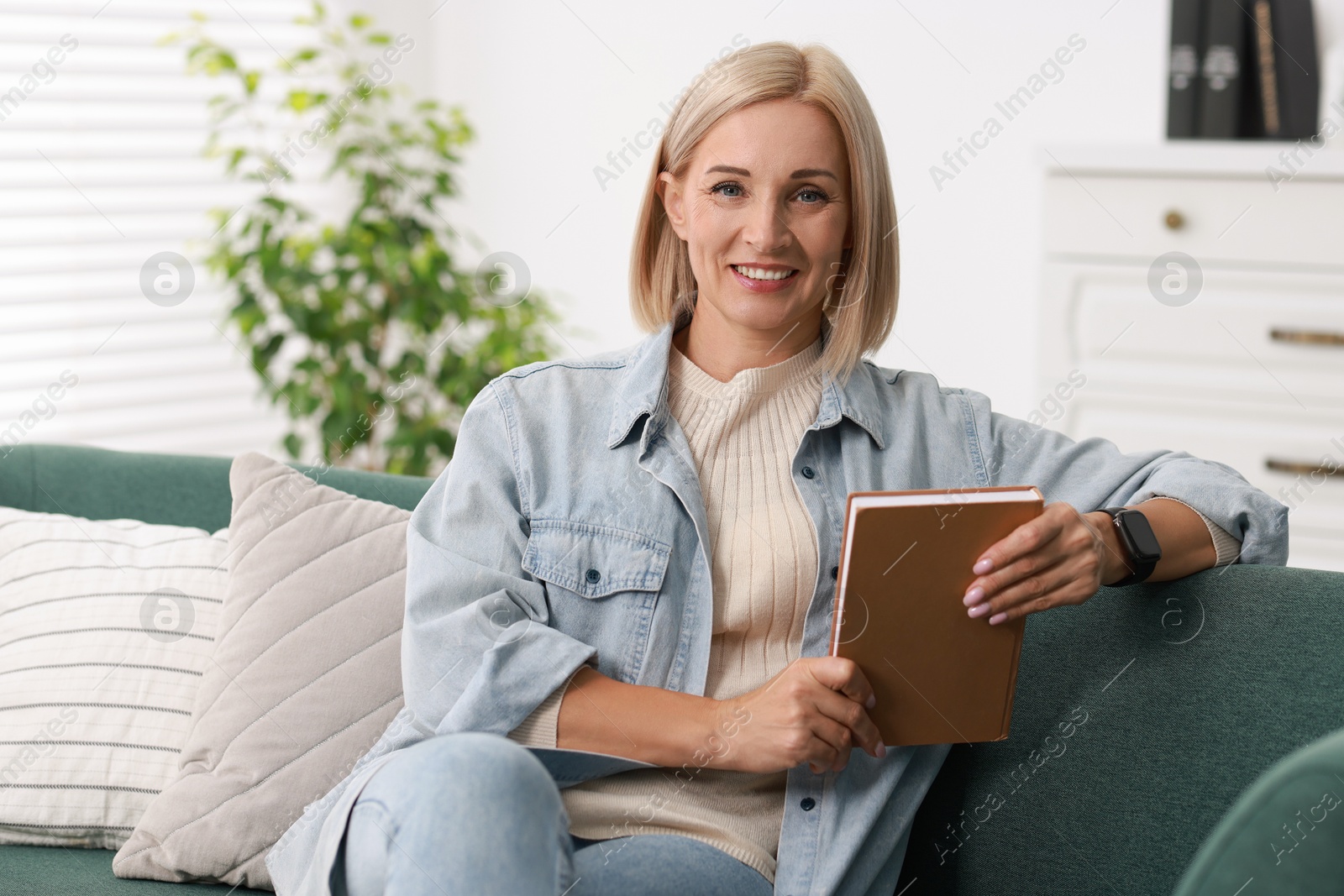 Photo of Portrait of smiling middle aged woman with book on sofa at home