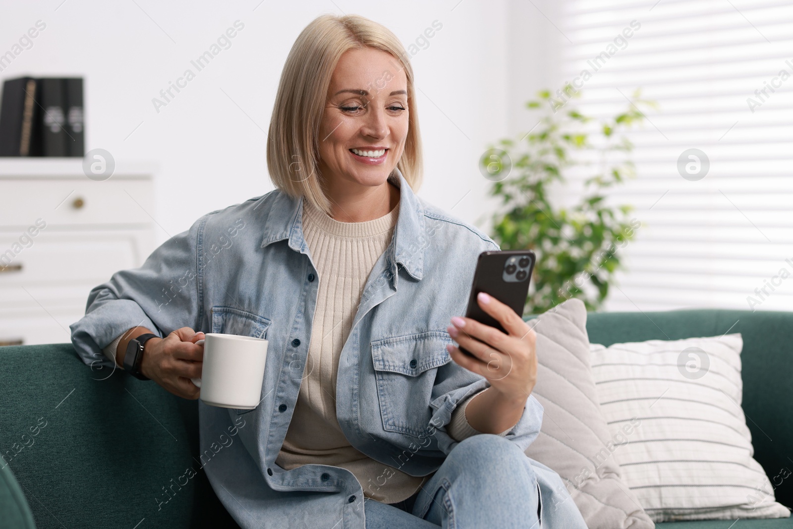 Photo of Smiling middle aged woman with cup of hot drink using smartphone at home