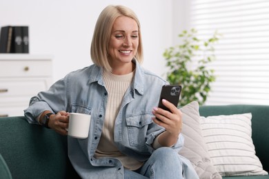 Photo of Smiling middle aged woman with cup of hot drink using smartphone at home