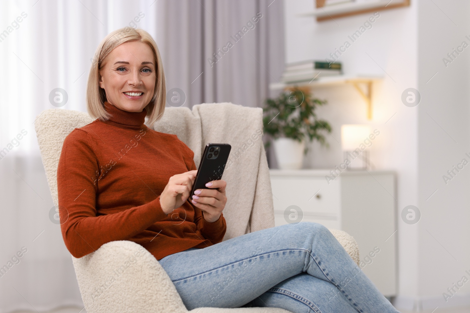 Photo of Portrait of smiling middle aged woman with smartphone on armchair at home