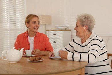 Caregiver and senior woman enjoying hot drink at table indoors