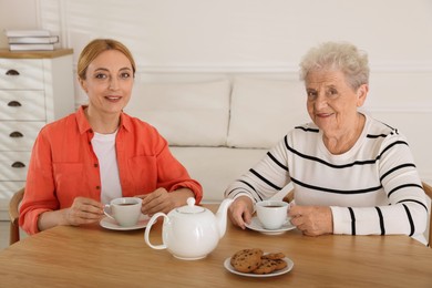 Caregiver and senior woman enjoying hot drink at table indoors