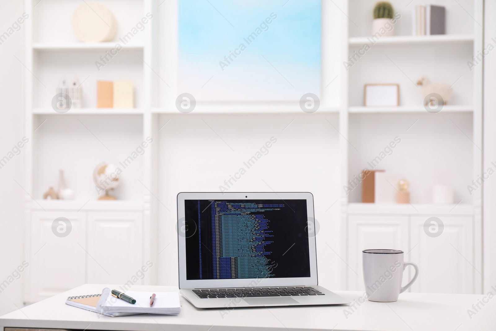 Photo of Programmer's workplace. Modern laptop with unfinished code, cup and stationery on white desk indoors