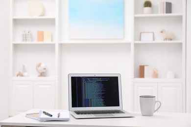 Photo of Programmer's workplace. Modern laptop with unfinished code, cup and stationery on white desk indoors