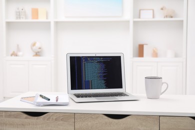 Photo of Programmer's workplace. Modern laptop with unfinished code, cup and stationery on desk indoors