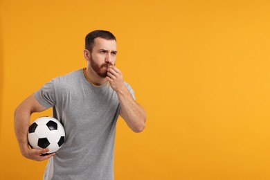 Young man with soccer ball blowing whistle on orange background, space for text