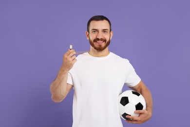 Happy young man with whistle and soccer ball on purple background