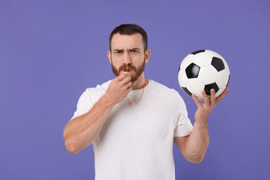 Young man with soccer ball blowing whistle on purple background