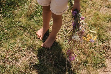 Photo of Little girl with bouquet of wildflowers walking barefoot outdoors on sunny day, closeup. Child enjoying beautiful nature