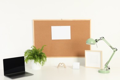 Photo of Cork board with blank paper note, laptop, lamp and decorative elements on light table near white wall