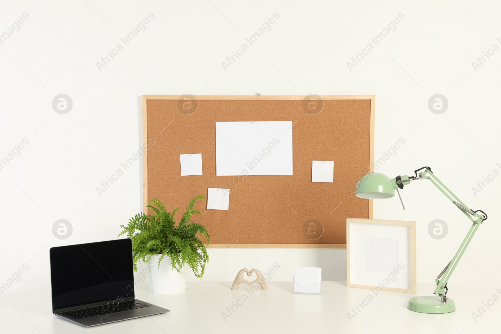 Photo of Cork board with blank paper notes, laptop, lamp and decorative elements on light table near white wall