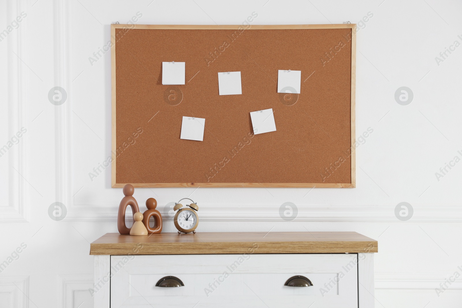 Photo of Cork board with blank paper notes, alarm clock and decorative figures on chest of drawers