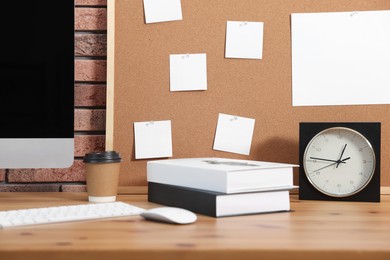 Cork board with blank paper notes, computer, books and alarm clock on wooden table