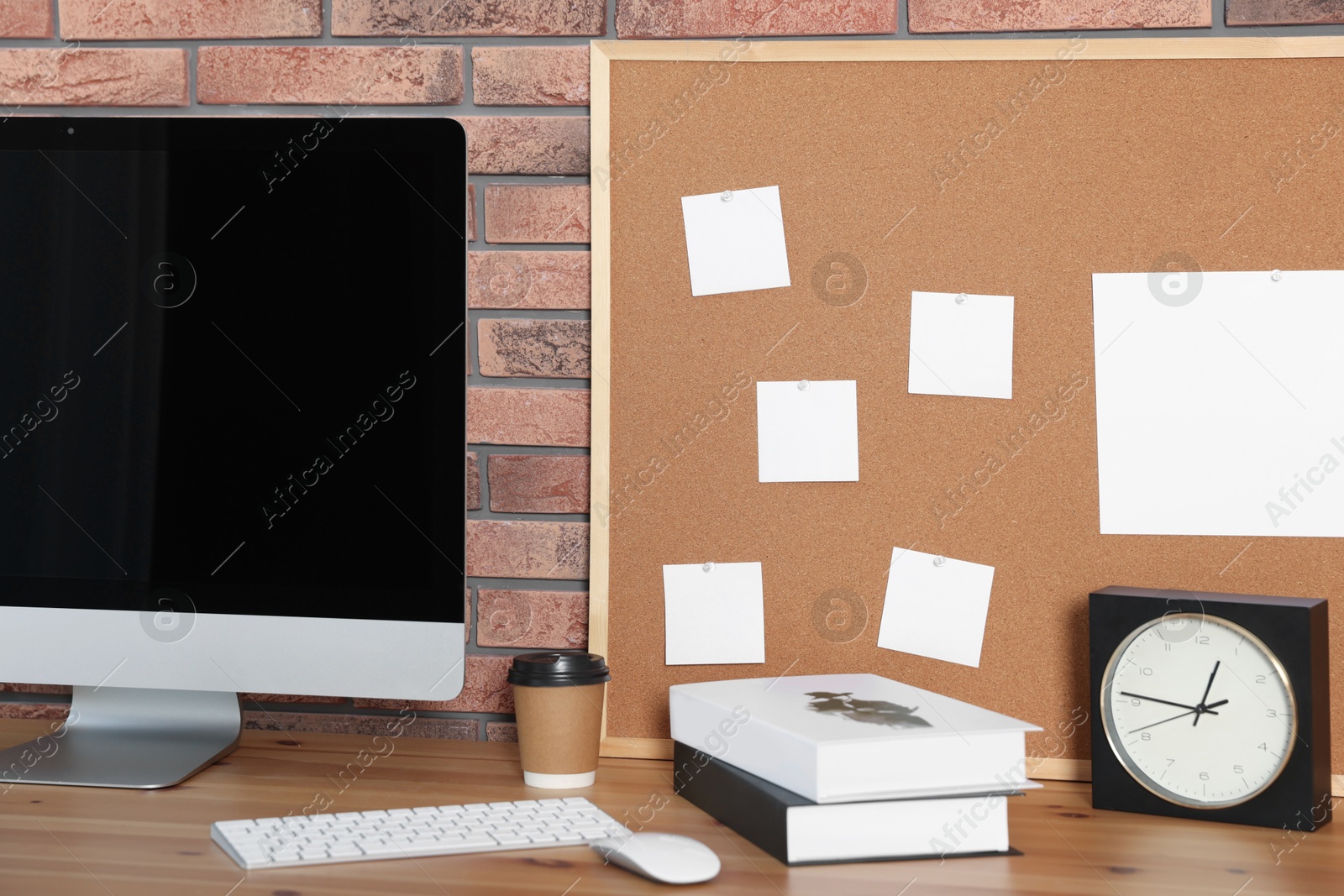 Photo of Cork board with blank paper notes, computer, books and alarm clock on wooden table near brick wall